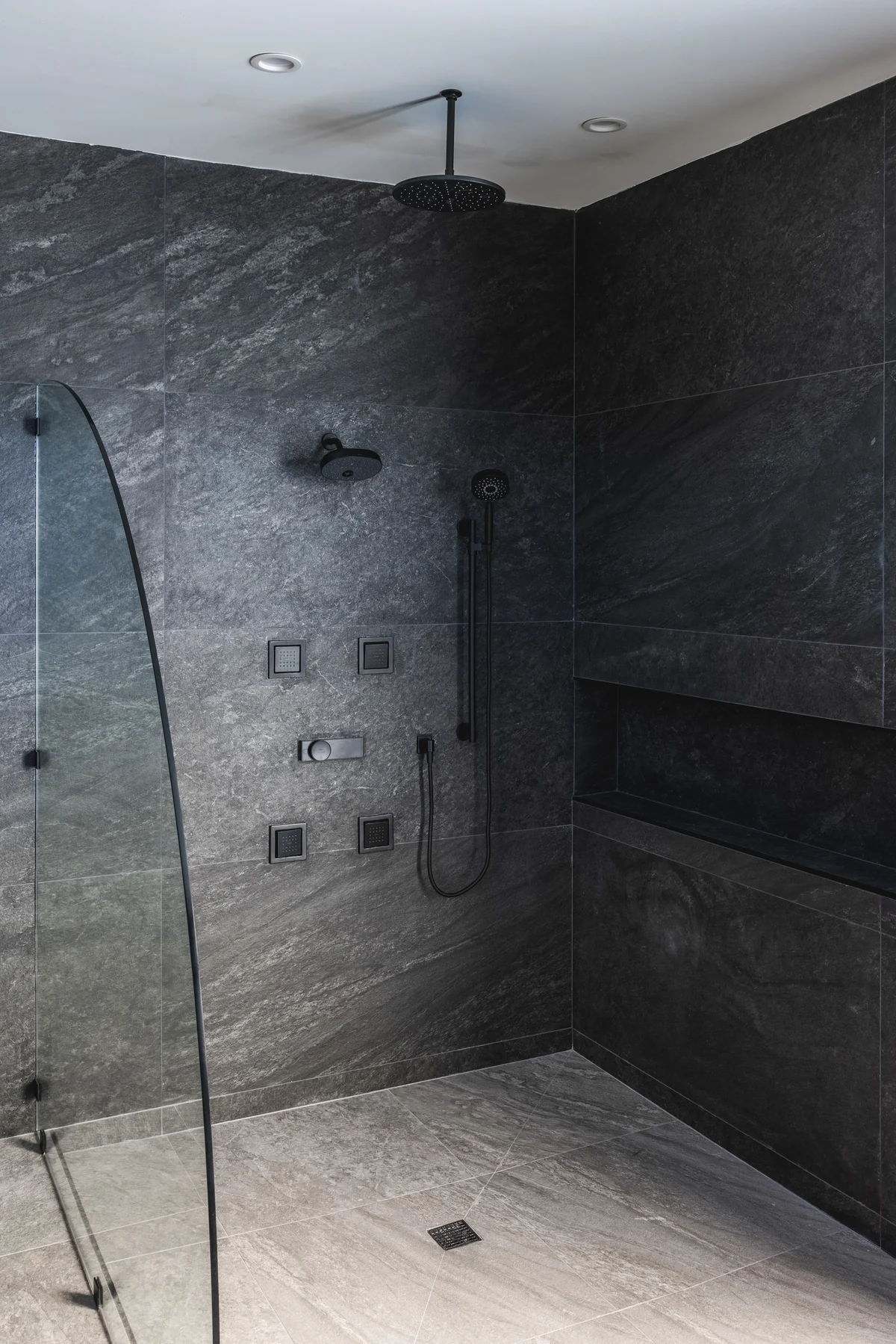 Walk-in shower with dark wall tile, overhead shower fitting, and recessed shelving